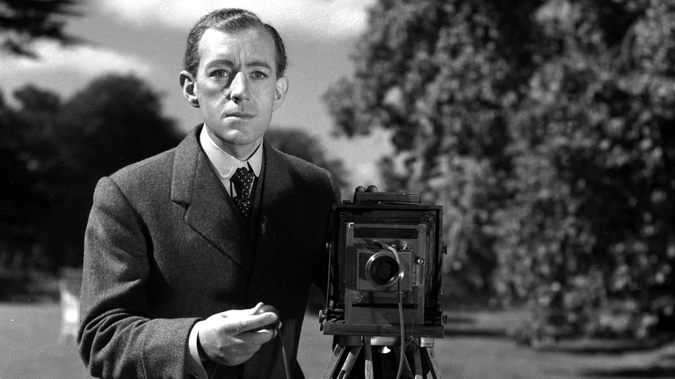 Alec Guiness' character is standing next an old frame camera, holding the lense clap looking into into the distance