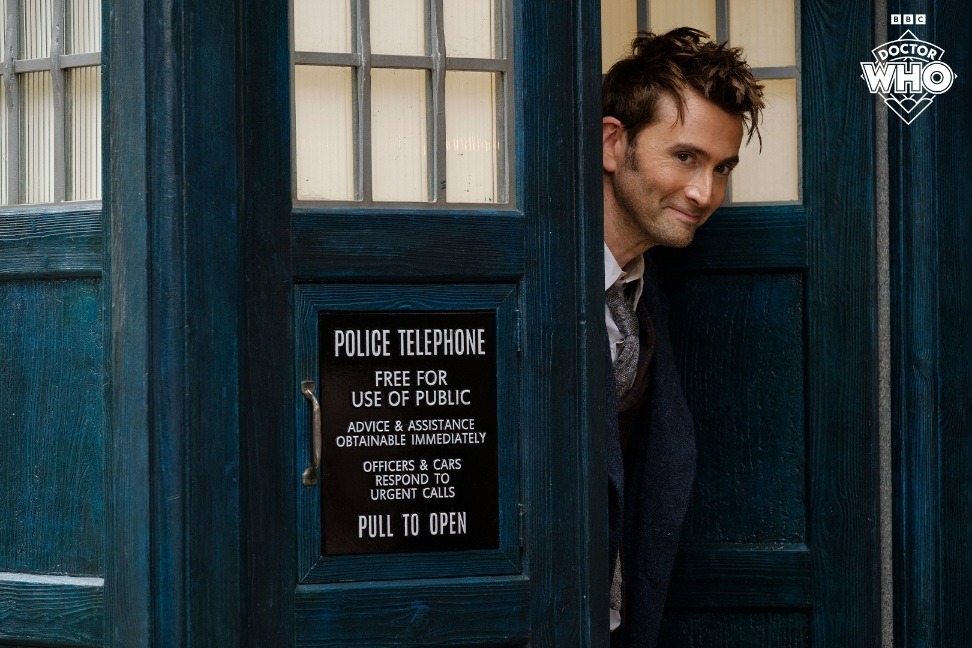 David Tennant as the Doctor, peering out from the TARDIS doors with a smile on his face in the upcoming 60th anniversary specials.