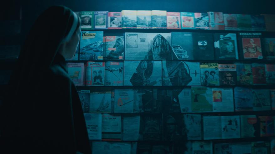 Taissa Farmiga dressed as a nun, standing infront of magazines that has a demon on them. Cool blue lighting