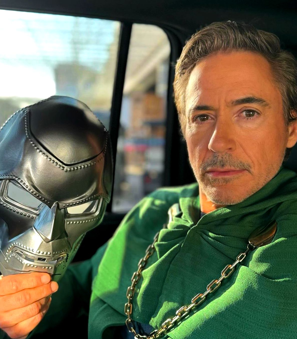 Actor Robert Downey Jr. Is sitting in a car wearing a green cloak and holding a Doctor Doom mask