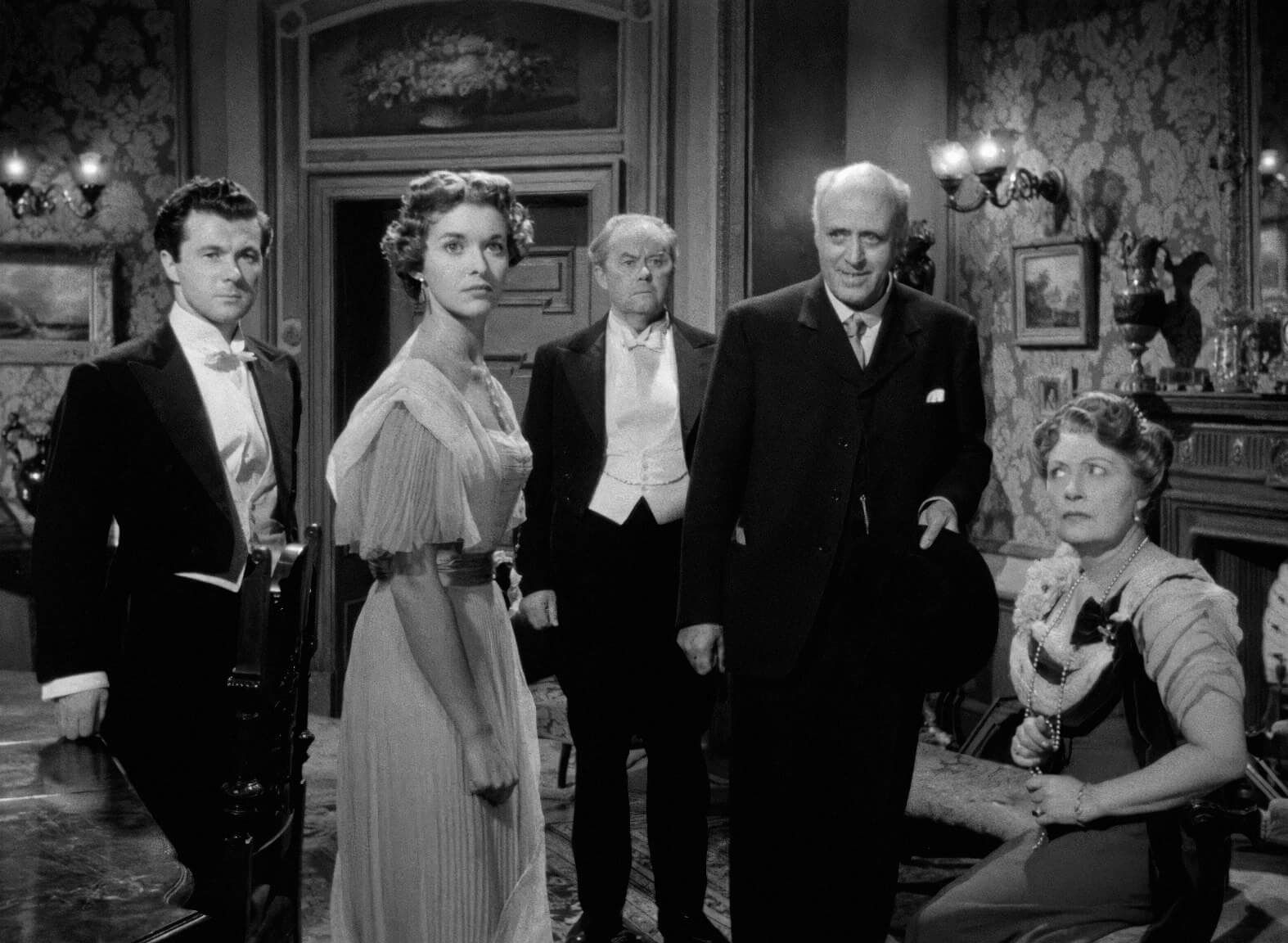 Well-dressed characters gather around in an evening parlour room scene, black and white image.