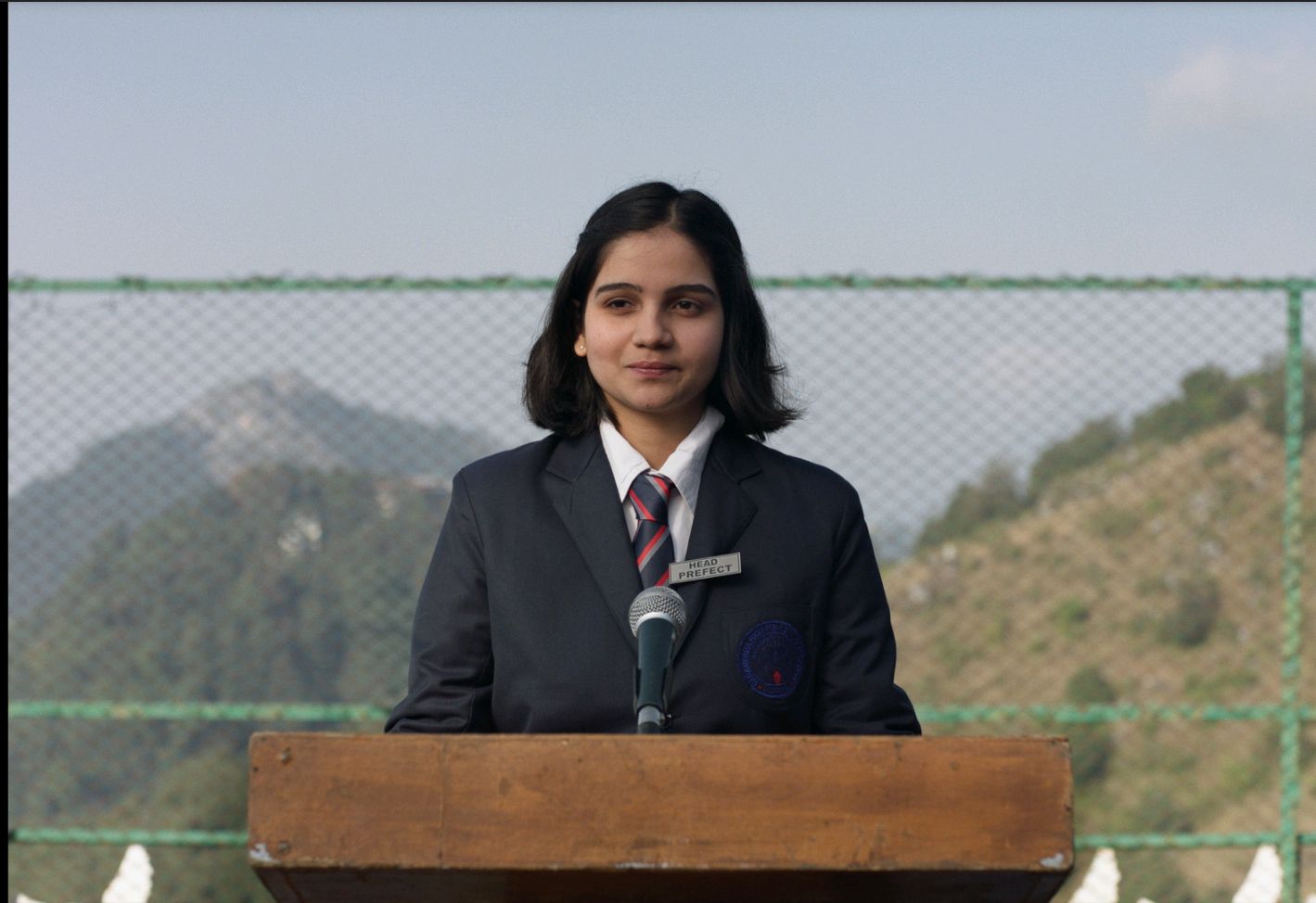 Mila is wearing a school uniform, standing on a podium as head prefect, in Girls Will Be Girls