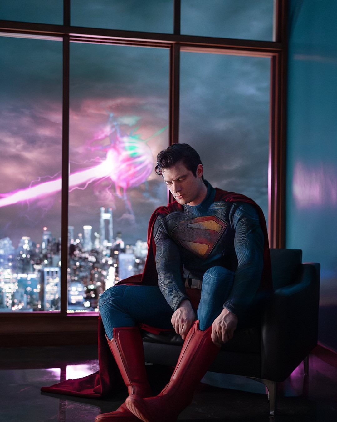 David Corenswet sitting in a chair, dressed in the Superman suit in a building with laser in the sky