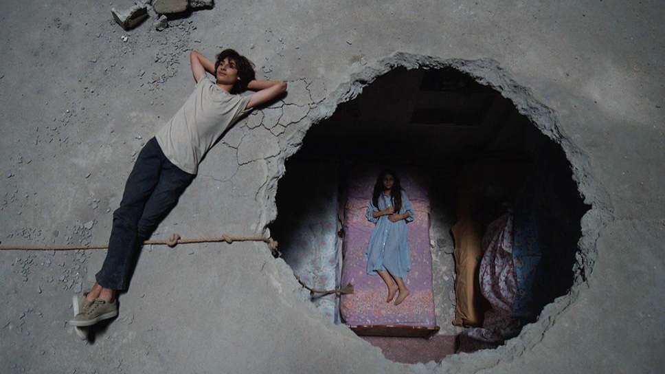 Fim promo image: Damascas building with a hole in it and someone lounging on the roof while a woman lays on a dusty bed in the hole caused by bomb