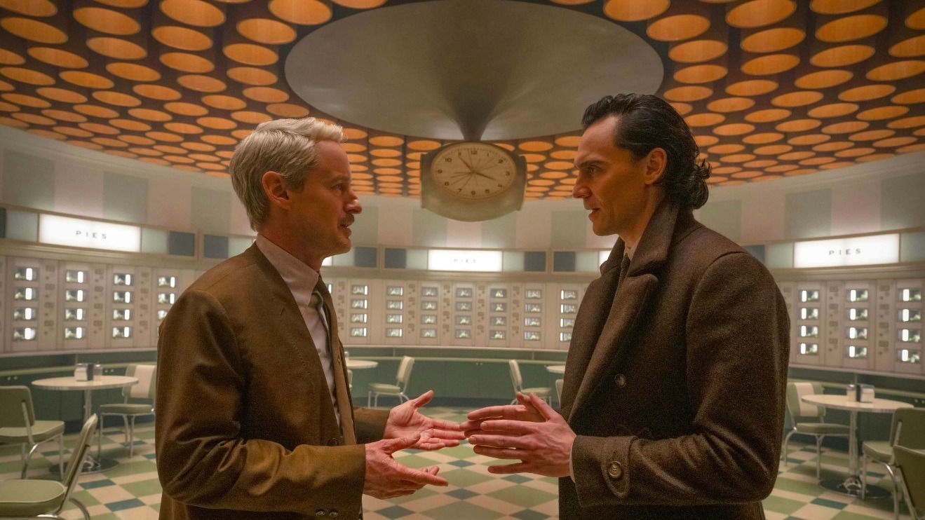 Mobius is talking to Loki, at the TVA. They are both wearing suits. Mobius has a brown suit jacket and Loki has a darker grey jacket. There's also a clock on the ceiling
