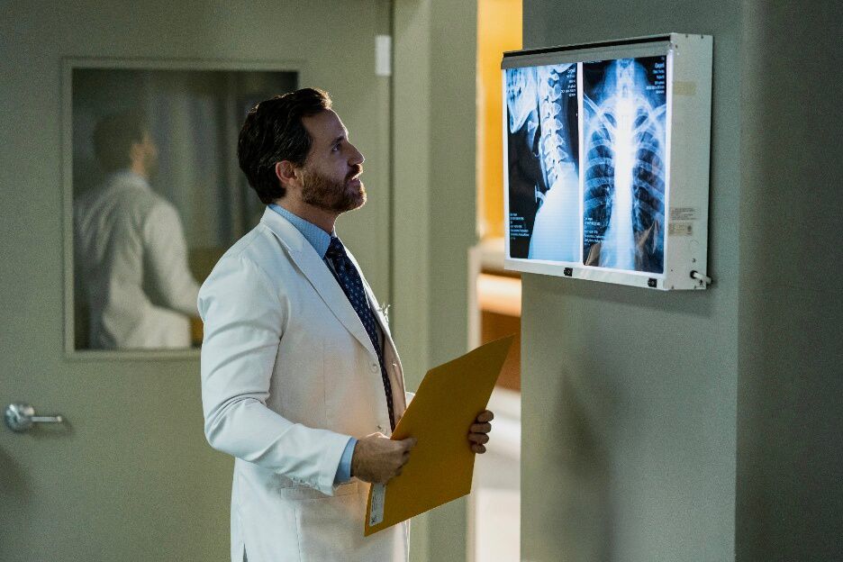 Edgar Ramírez is in white Doctors coat looking at an X-Ray scan of a person spine and ribs in Dr Death