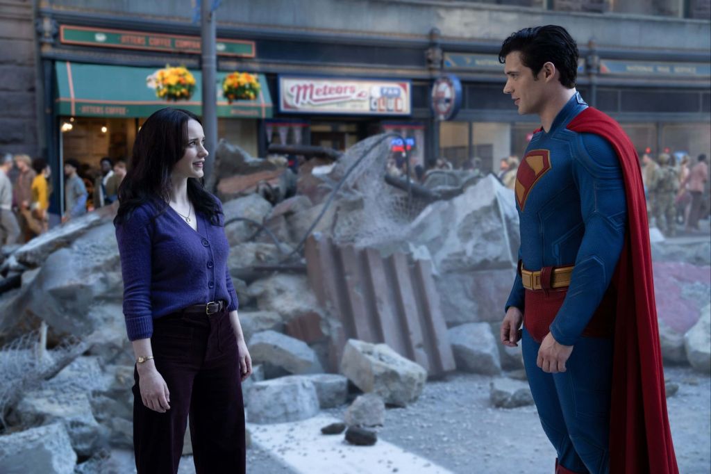 Lois Lane is standing in front of rubble meeting Superman.